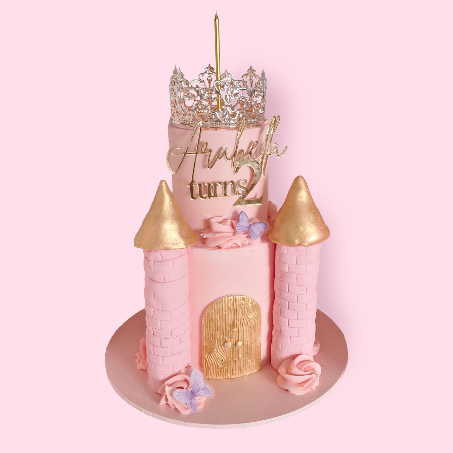 Pink princess castle-themed birthday cake with gold turrets, butterflies, and silver crown topper, personalised for Arabella turning 2