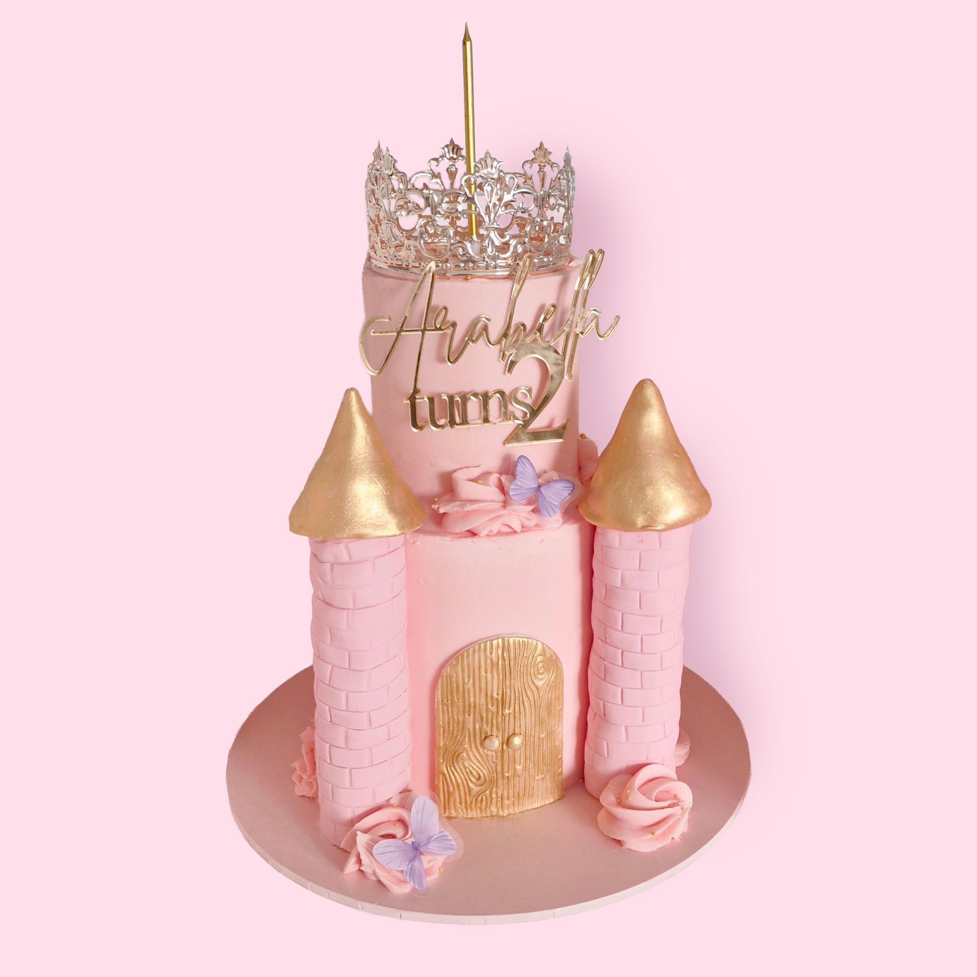 Pink princess castle-themed birthday cake with gold turrets, butterflies, and silver crown topper, personalised for Arabella turning 2